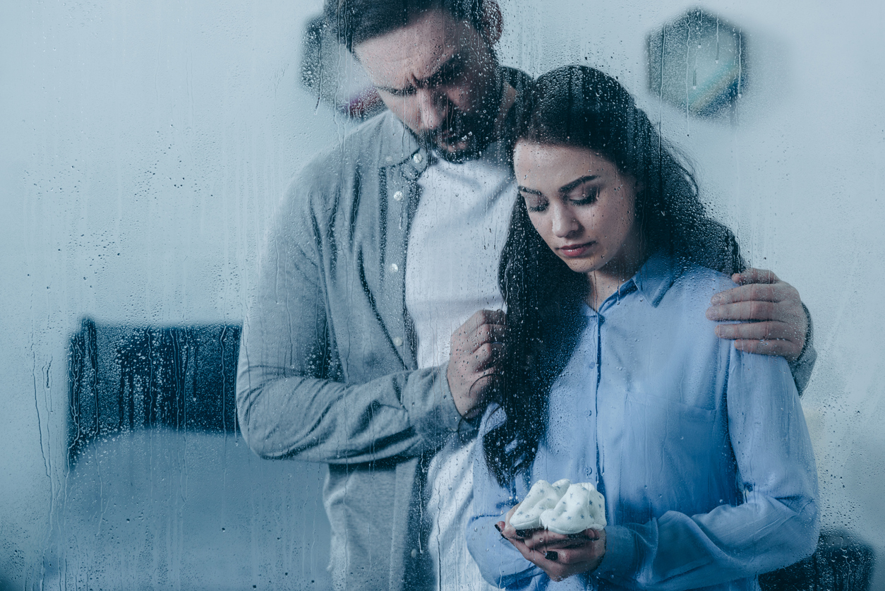 husband grieving and hugging wife with baby shoes through window with raindrops